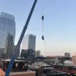 Crane lifting a rooftop unit beside a brick building, with a white truck below